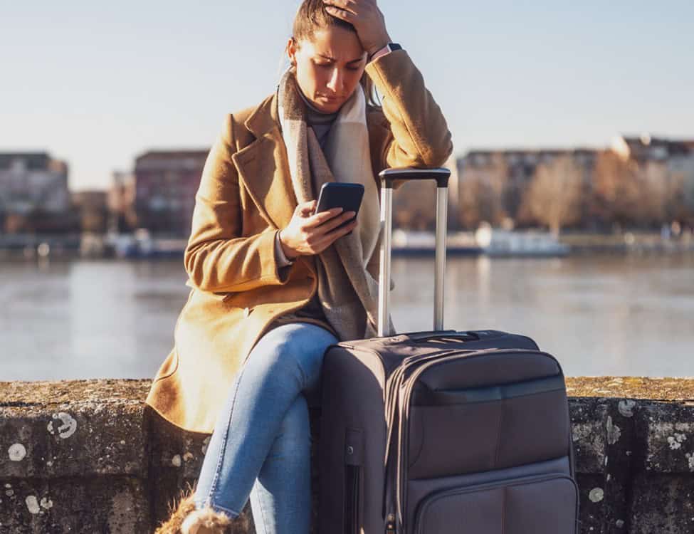 Image of a female traveller, sitting on a harbour wall, looking at her phone and looking distressed.